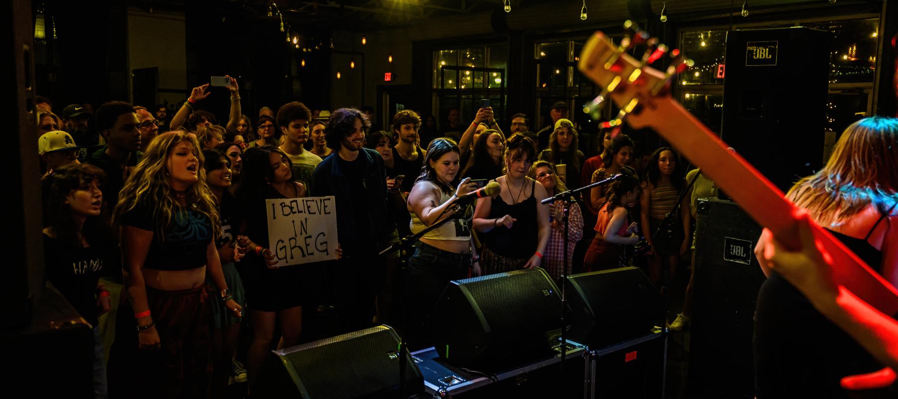 Shot of the crowd at a concert. Some are singing along, some are recording the performace and someone is holding a sign.